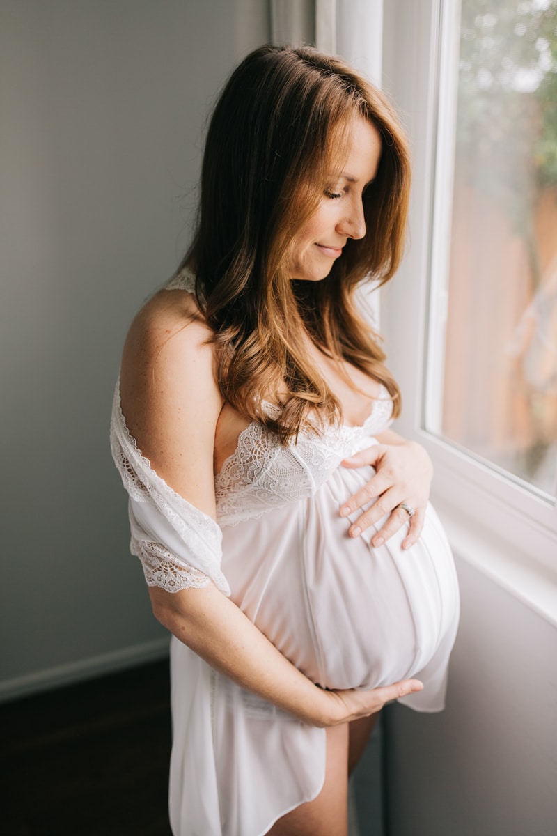 Maternity Photographer, woman in white dress standing next to a window