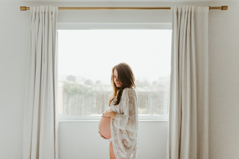 Maternity Photographer, woman in lace cover up standing next to a window