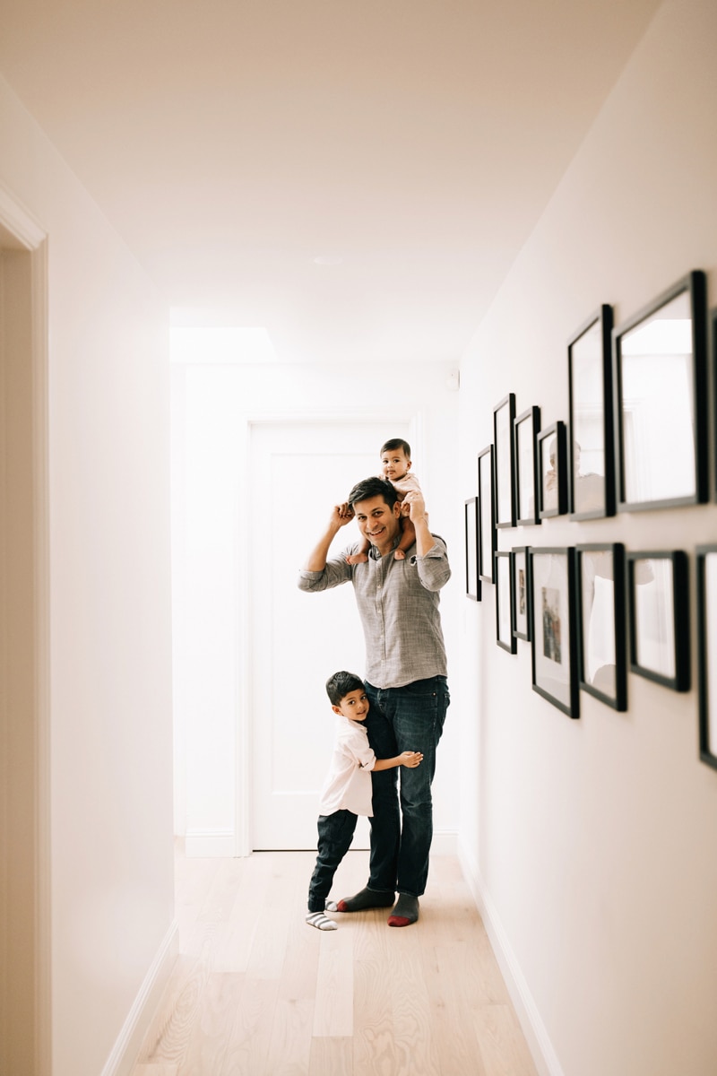 Family Photographer, dad standing in hallway with baby on his shoulders and little boy hugging his leg